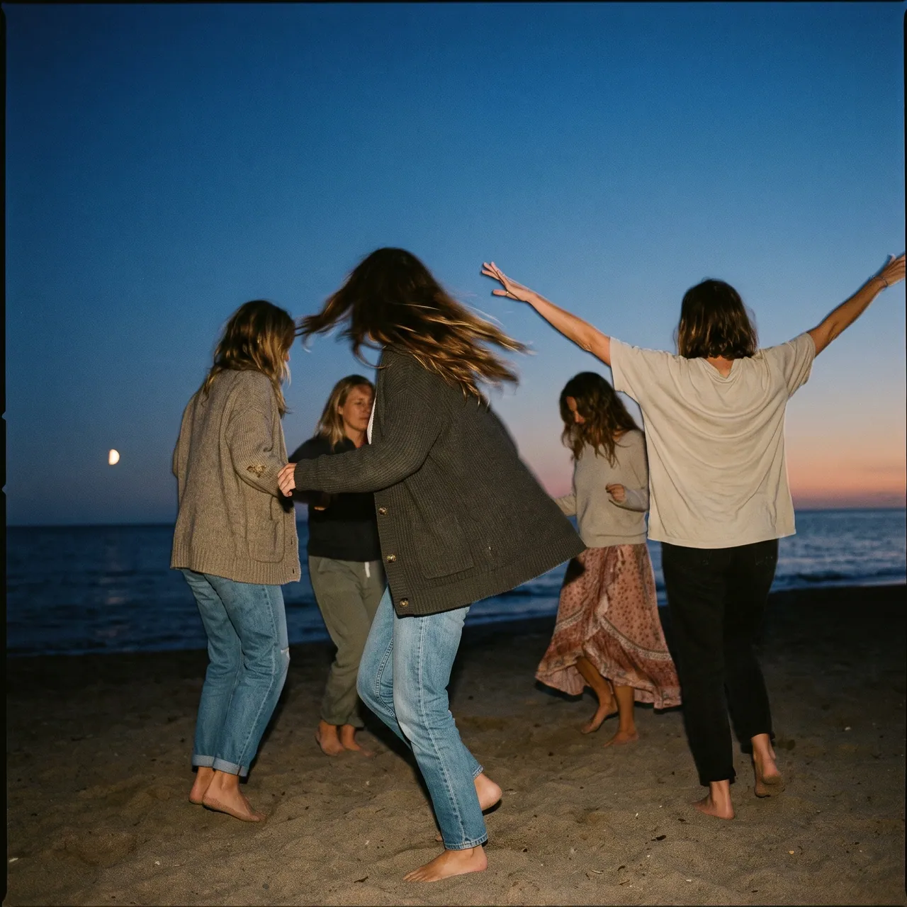 Cinco mujeres bailan descalzas en la playa al anochecer, con una luna pequeña sobre el mar